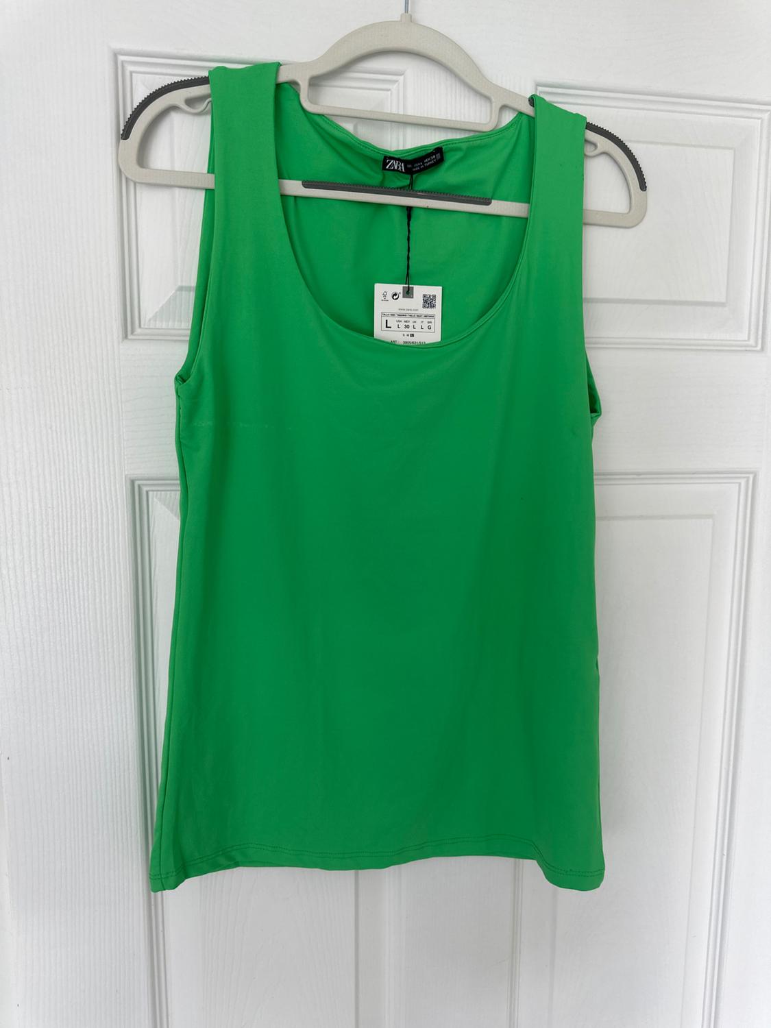 Green sleeveless t-shirt hanging on a grey coat hanger in front of a white door. Label visible with size details and QR code.