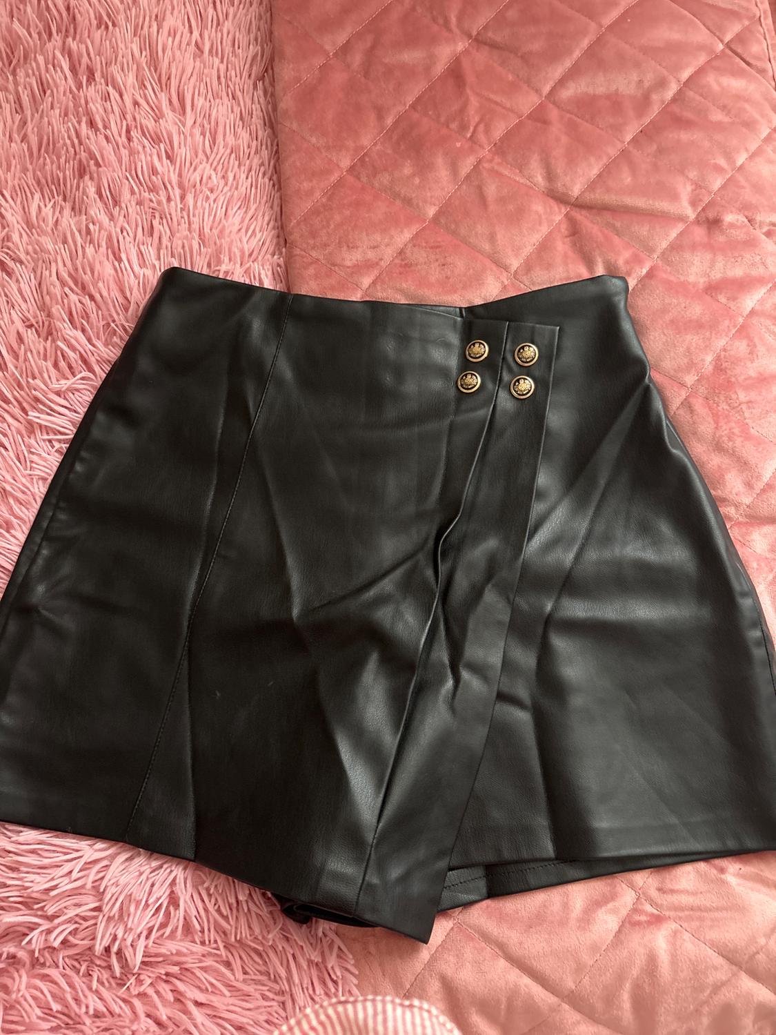 Black short skirt made of synthetic material with gold buttons, on a pink quilted bedspread and pink fluffy rug.