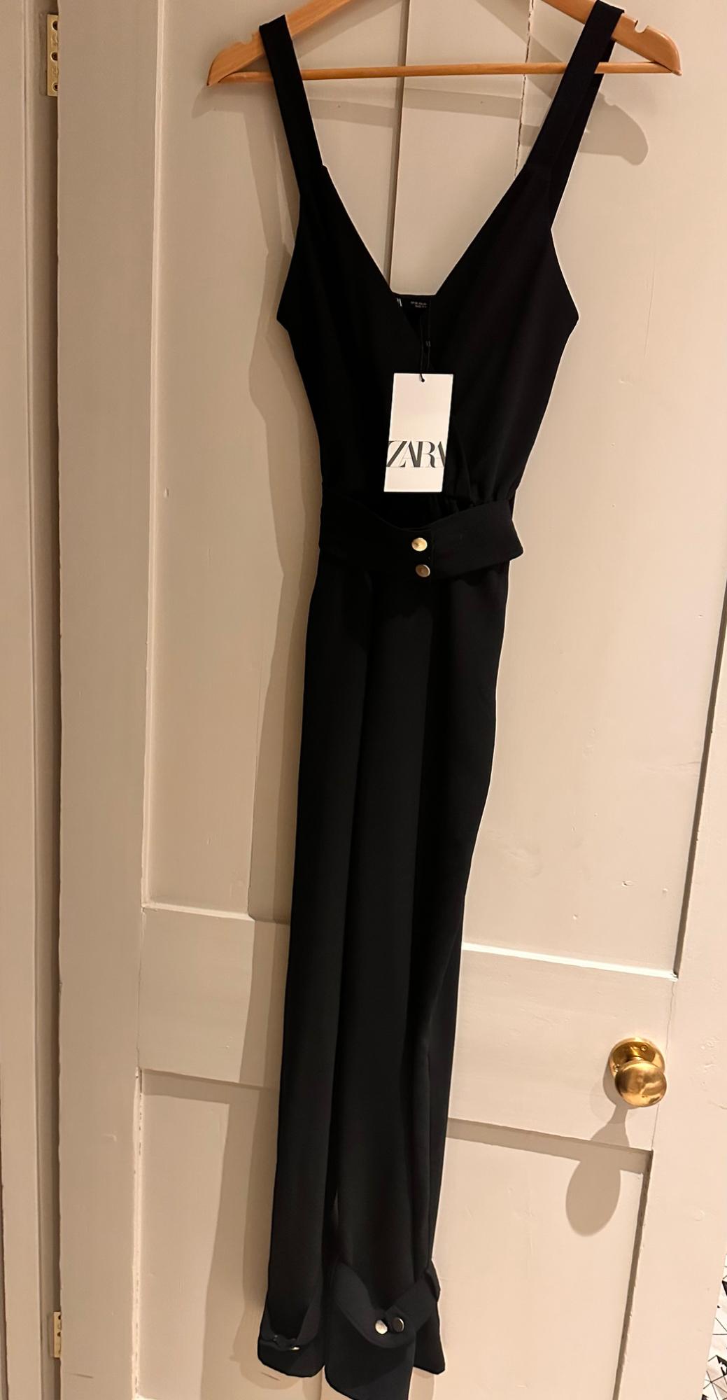 Sleeveless black jumpsuit hanging on a wooden hanger, with a visible label that reads "ZARA", over a white door.