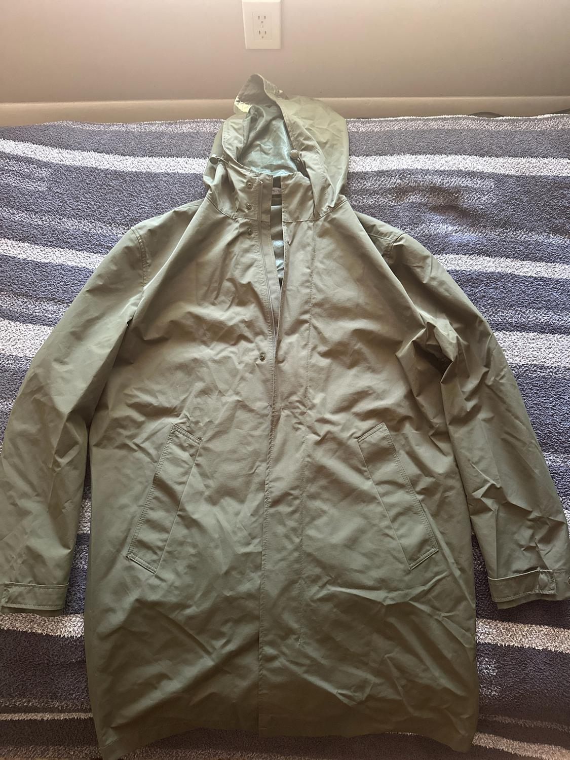 Olive green hooded jacket with pockets, spread out over a bed with a gray-and-white striped blanket.