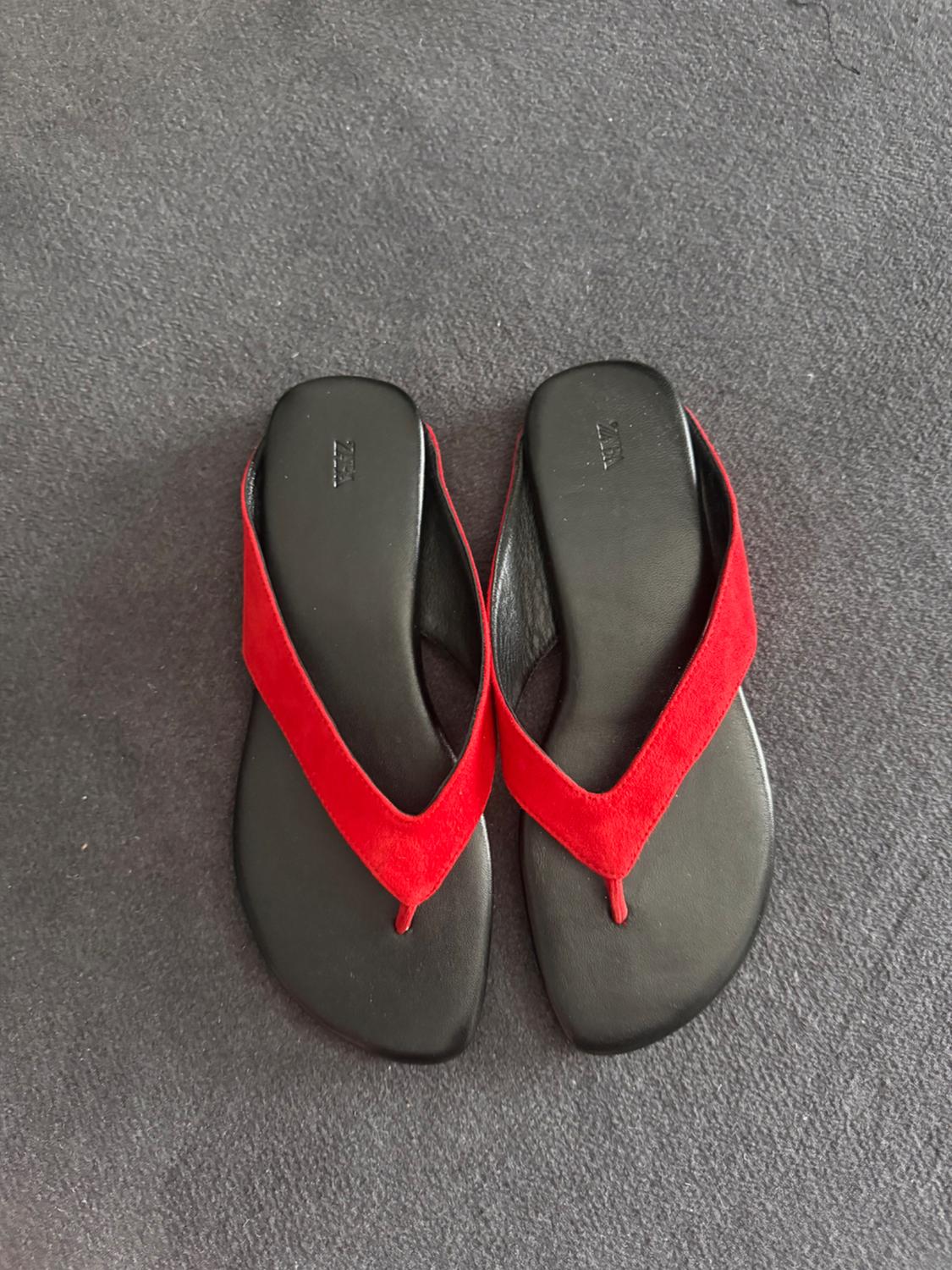 Pair of red thong flat sandals with a black sole on a textured grey surface. ZARA text on the insoles.