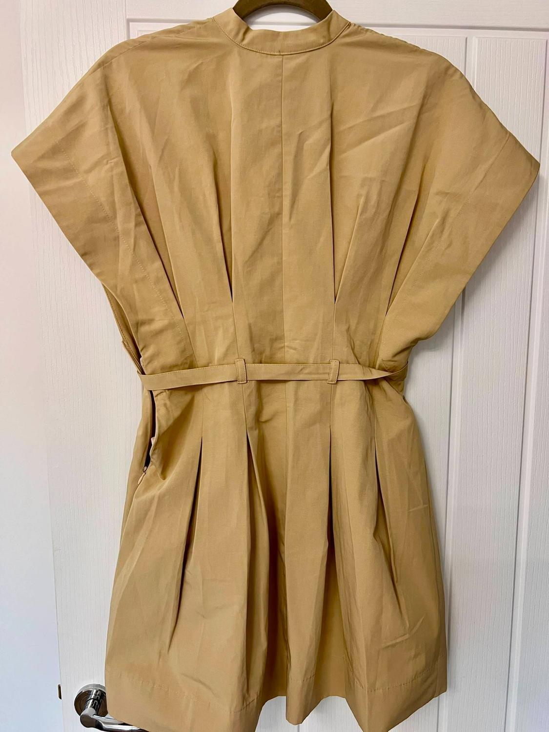 Beige dress with loose short sleeves, high neck, thin belt and pleated design, hanging on a hanger in front of a white door.