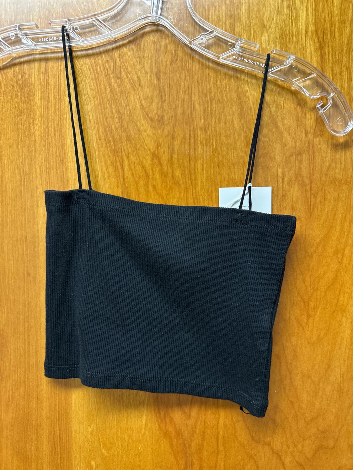 Black ribbed crop top with thin straps and label, hanging on a transparent hanger against a wooden background.