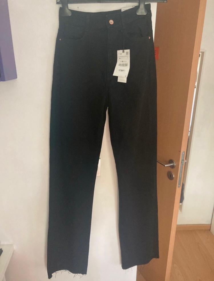 Black trousers hanging on a hanger, with a visible tag. Wooden door and white wall in the background.