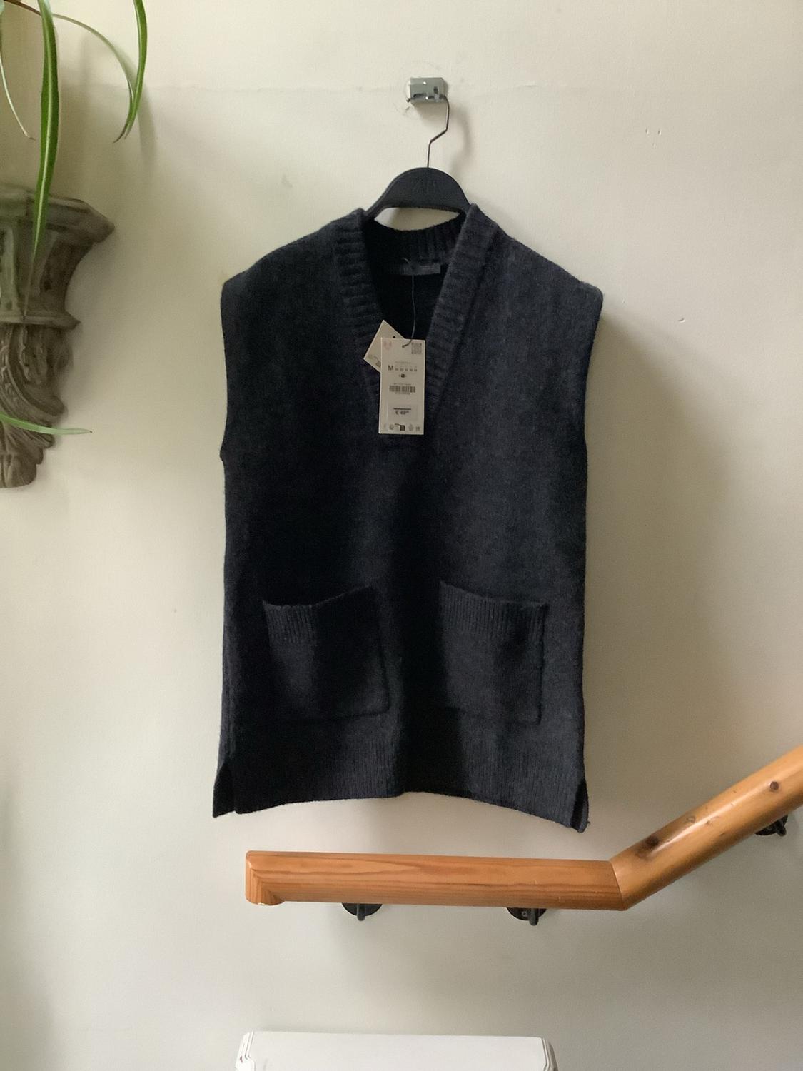 Dark grey knitted waistcoat with label, V-neck and pockets, hanging on a white wall next to a wooden handrail and a plant.