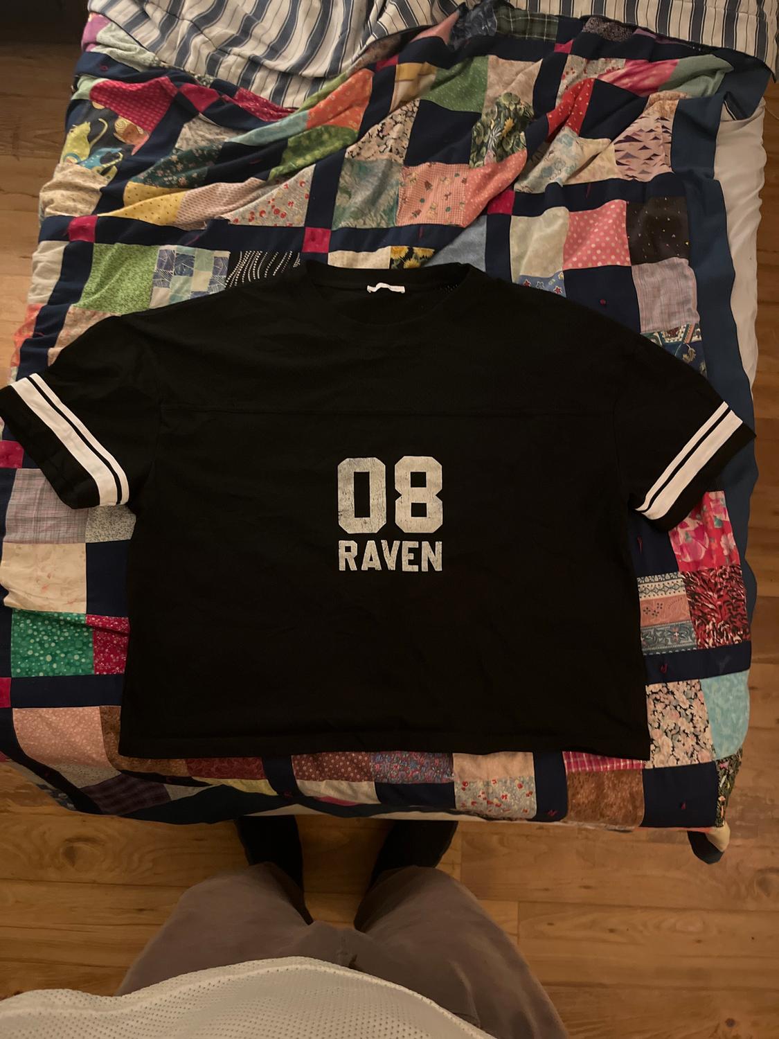 Black t-shirt with "08 RAVEN" text on a colourful patchwork quilt on a bed. Person standing at the edge.