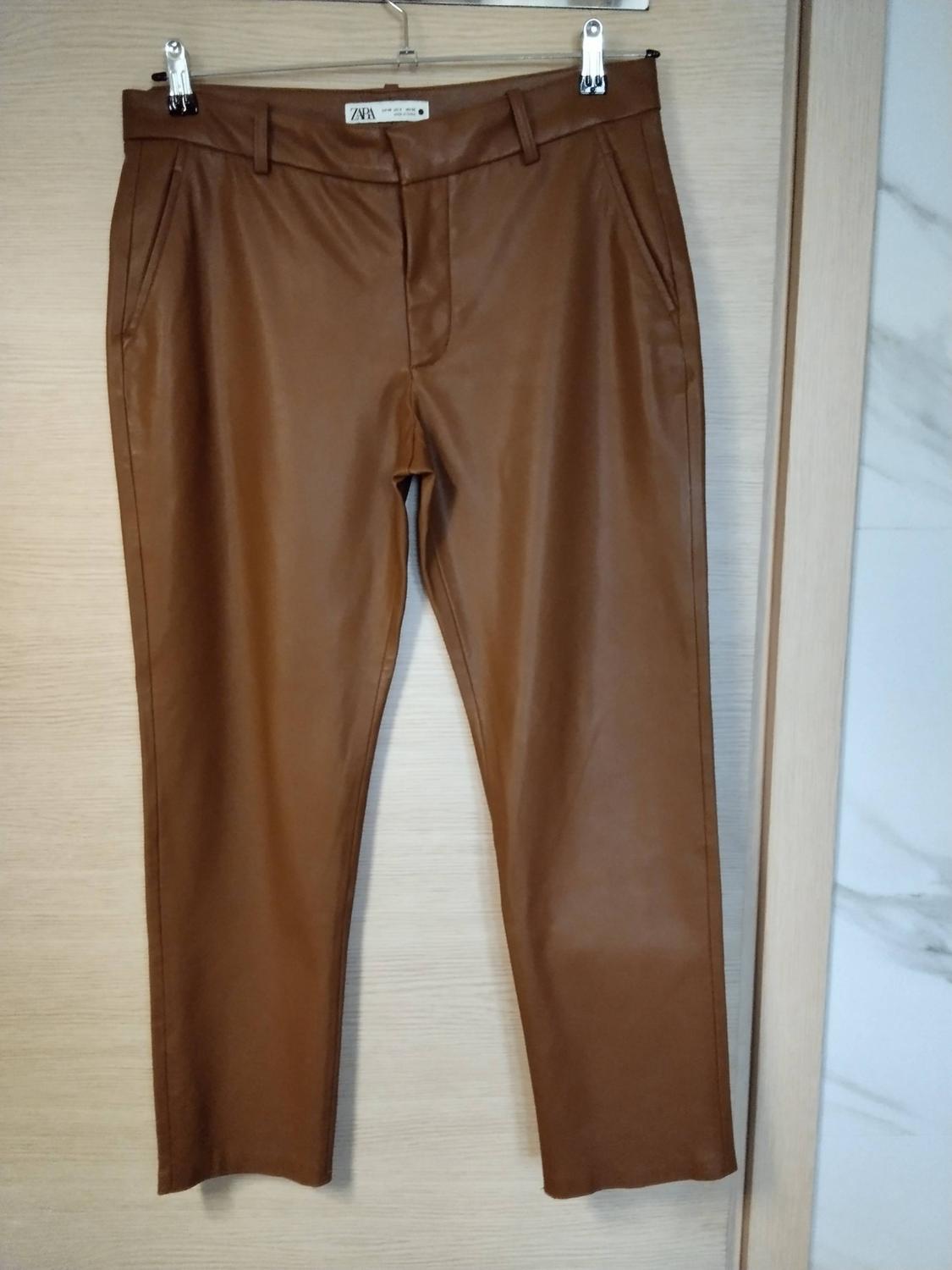 Brown trousers hanging on a metal hanger over a wooden door. The inside label reads ZARA.