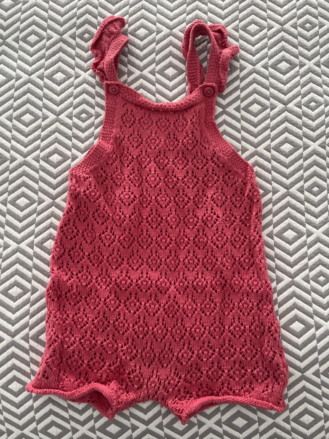 Pink knitted romper with straps and buttons, on a grey and white geometric patterned quilt.