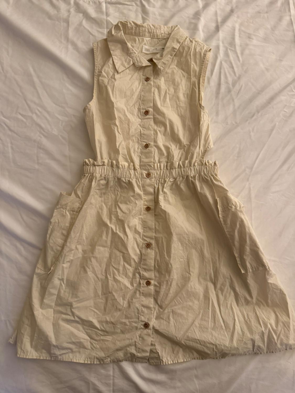 Front view of a sleeveless beige dress with a central button closure and side pockets.