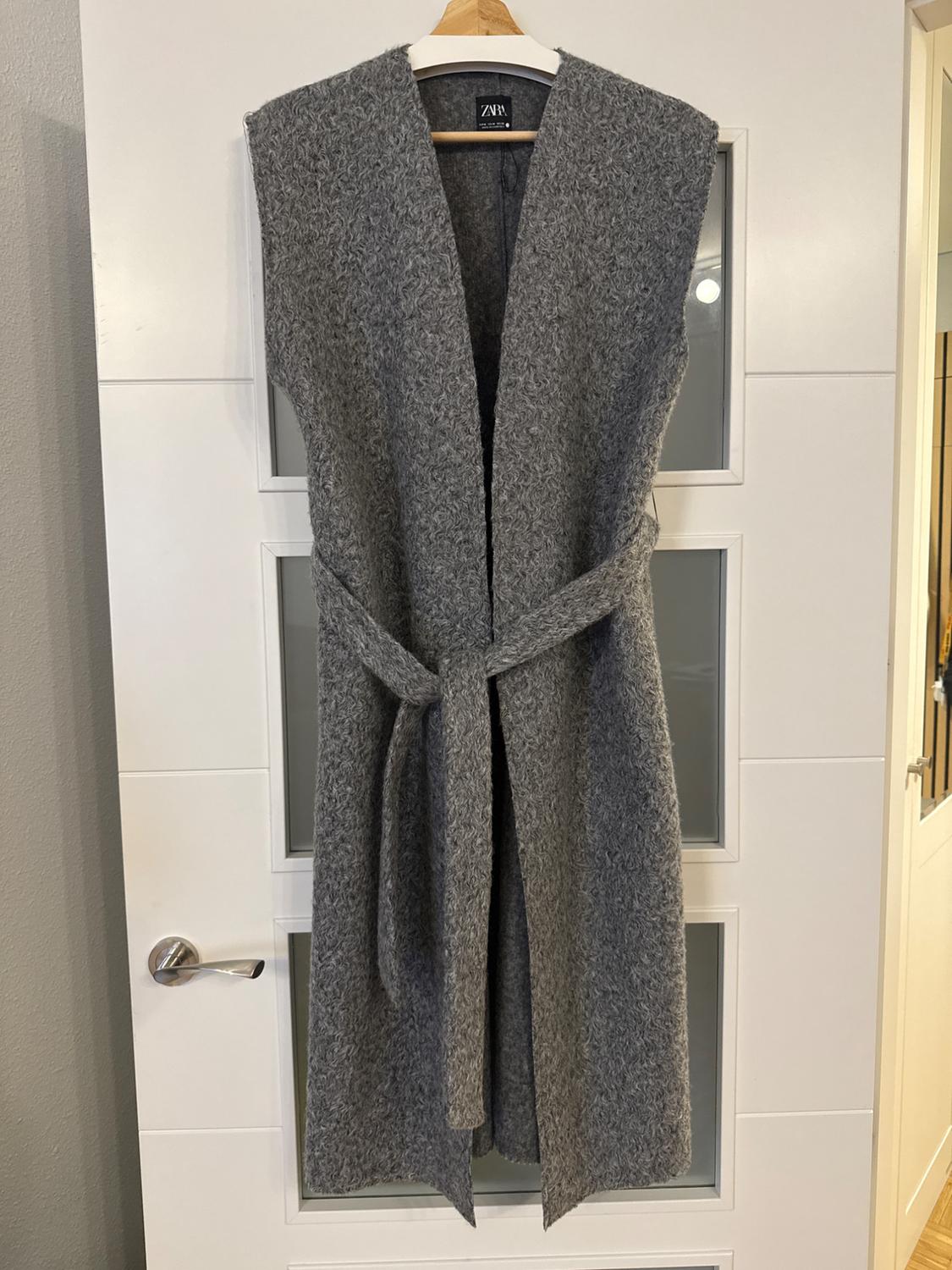 Textured grey longline waistcoat with a tie belt, hanging on a hanger over a white door. Label with the text ZARA.