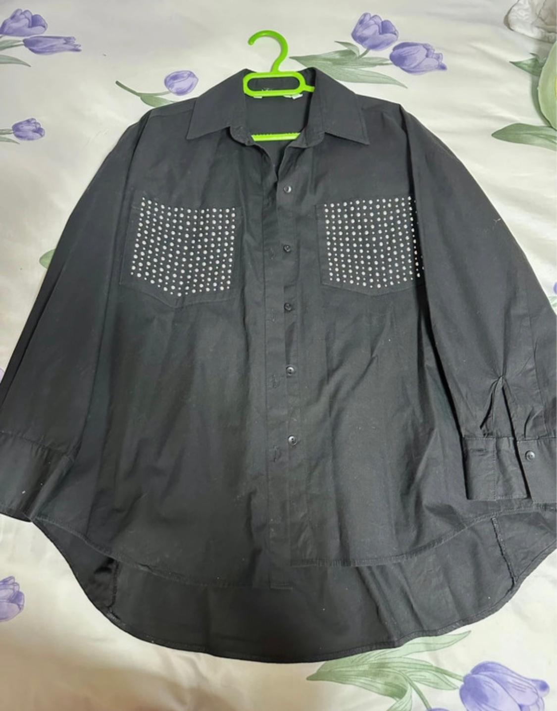 Black shirt with silver stud-embellished pockets, hanging on a green hanger over a bed with floral sheets.