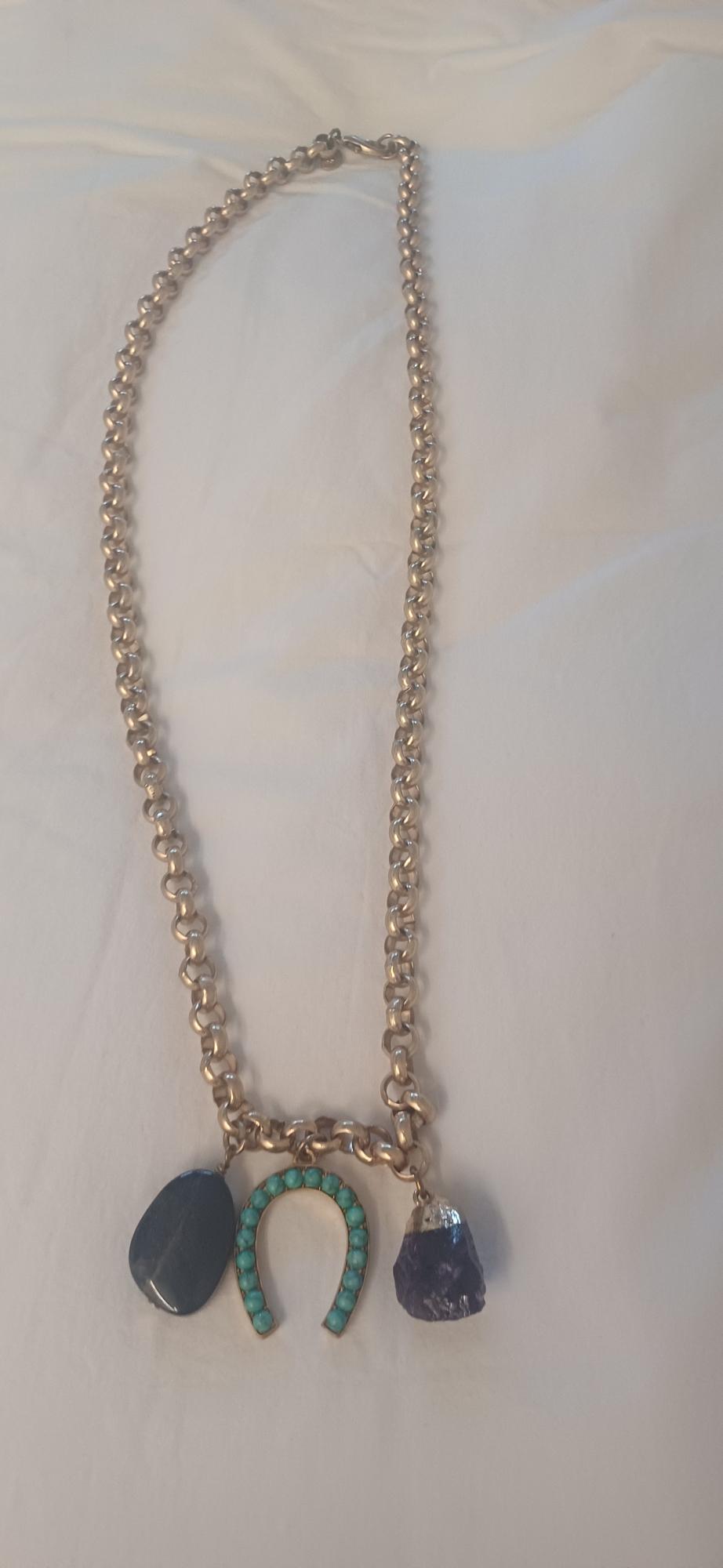 Gold chain necklace on white fabric with three pendants: a dark stone, a horseshoe with turquoises, and a purple stone.