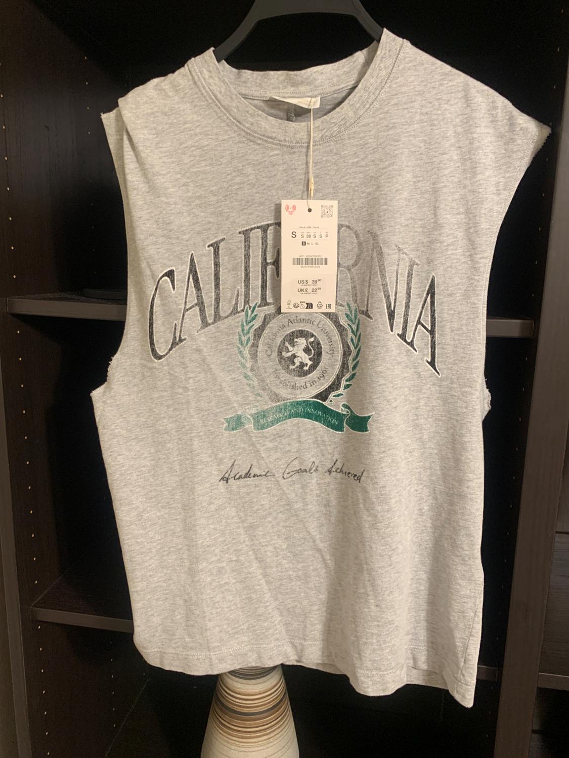 Grey sleeveless t-shirt with "CALIFORNIA" and "Academic Goals Achieved" text. Hanging price tag. Dark bookshelf background.