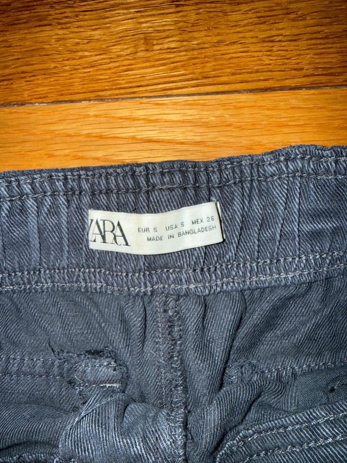 Zara pants tag in gray fabric, showing sizes EUR S, USA S, MEX 26, made in Bangladesh, on a wooden background.
