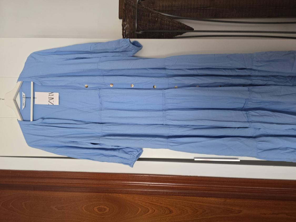 Light blue dress with gold buttons hanging on a white hanger. Visible label with the text "ZARA". White wall background.