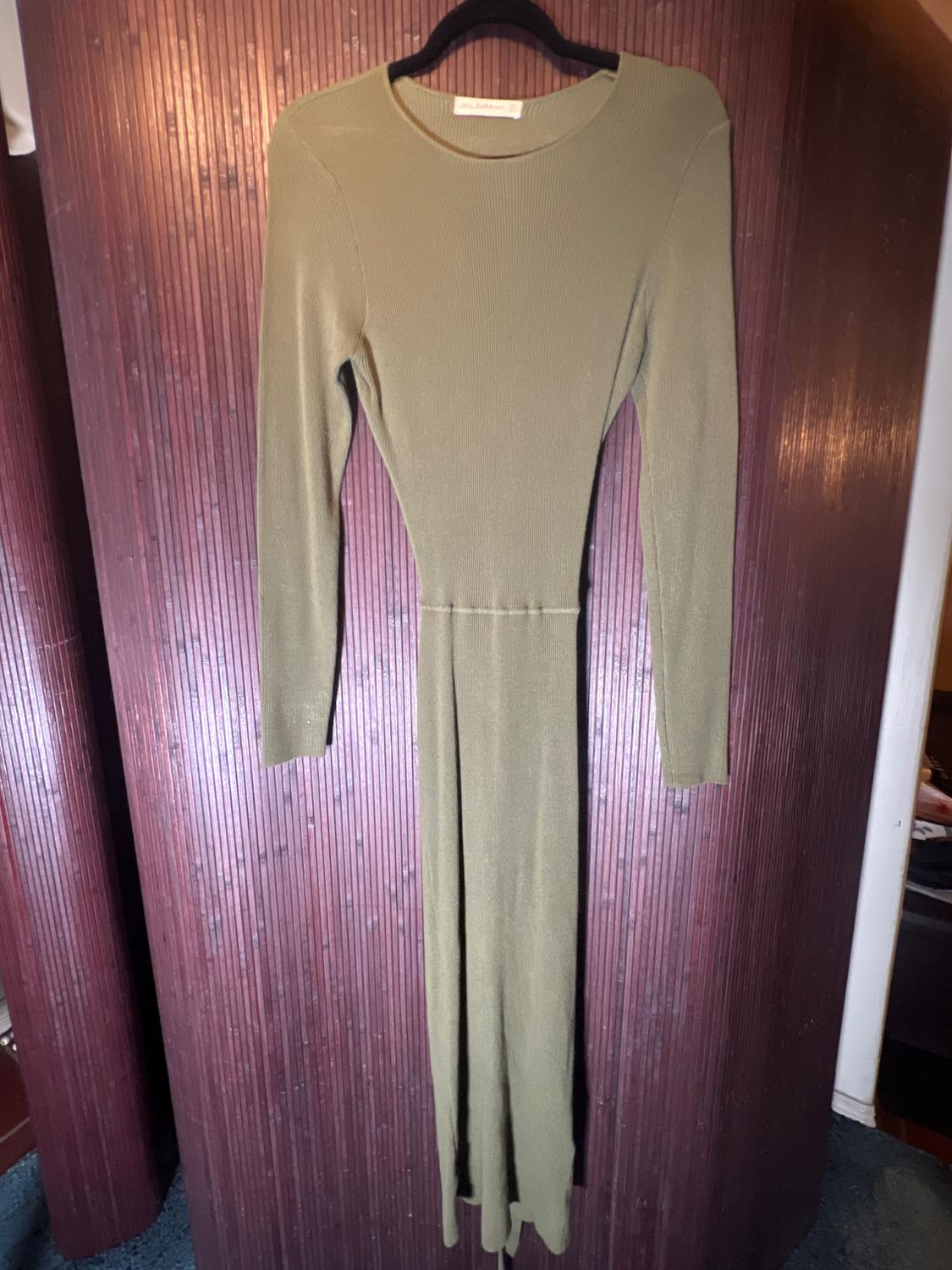 Olive green long-sleeved long dress hanging on a black hanger, against a background of brown wood panels.