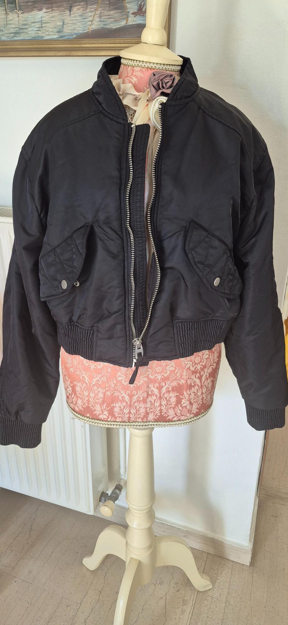 Mannequin wearing a black bomber jacket, with an open zip and flap pockets. Background with a white wall and a framed picture.