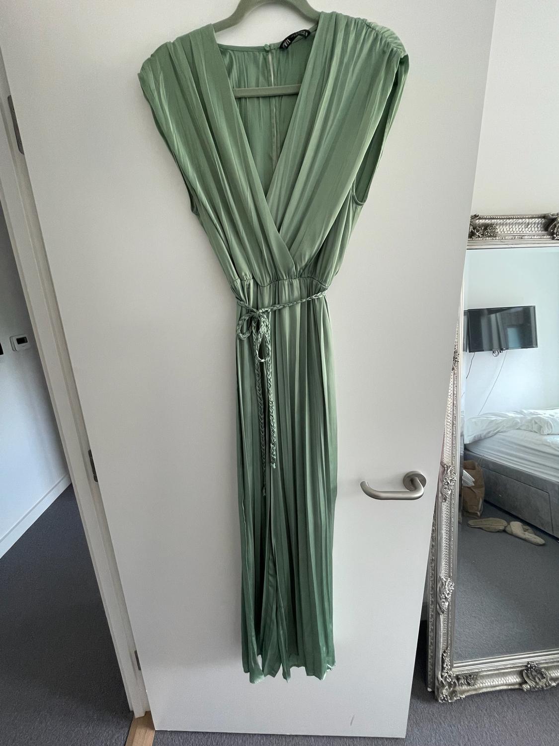 Light green pleated maxi dress with a wrap neckline and braided belt, hanging on a white door next to a mirror.