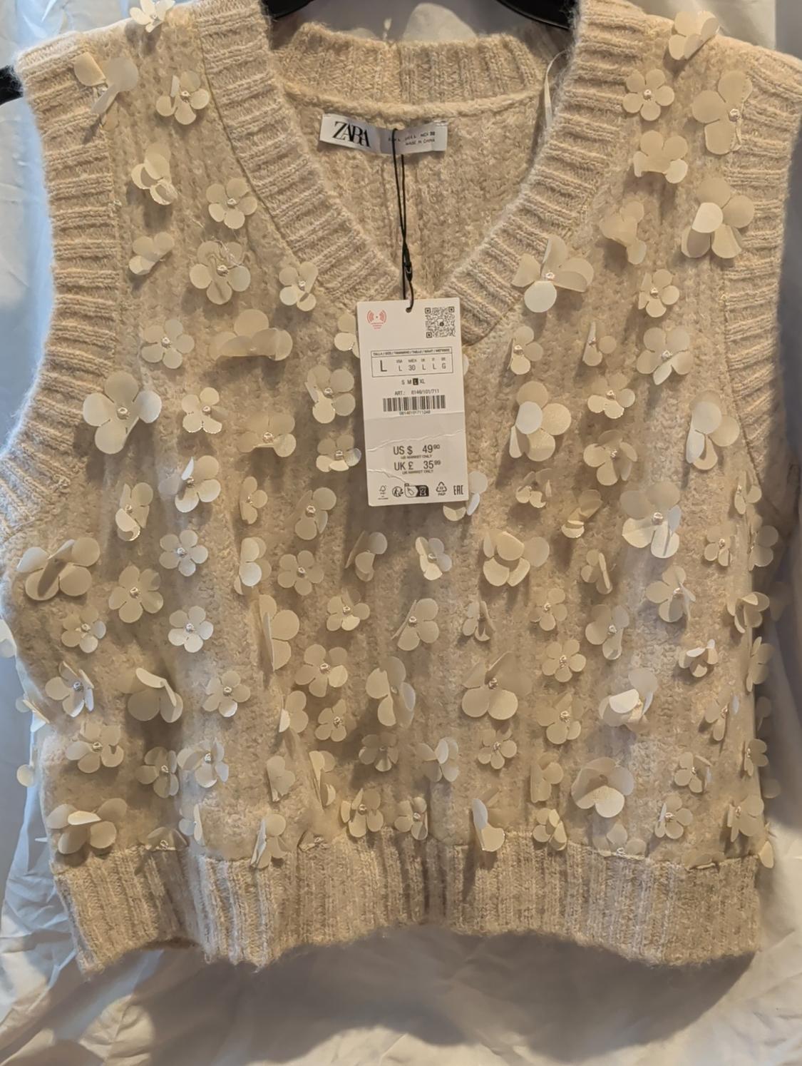Beige vest with embossed flower appliques, hang tag with $49.99 price, hanging on a white background.