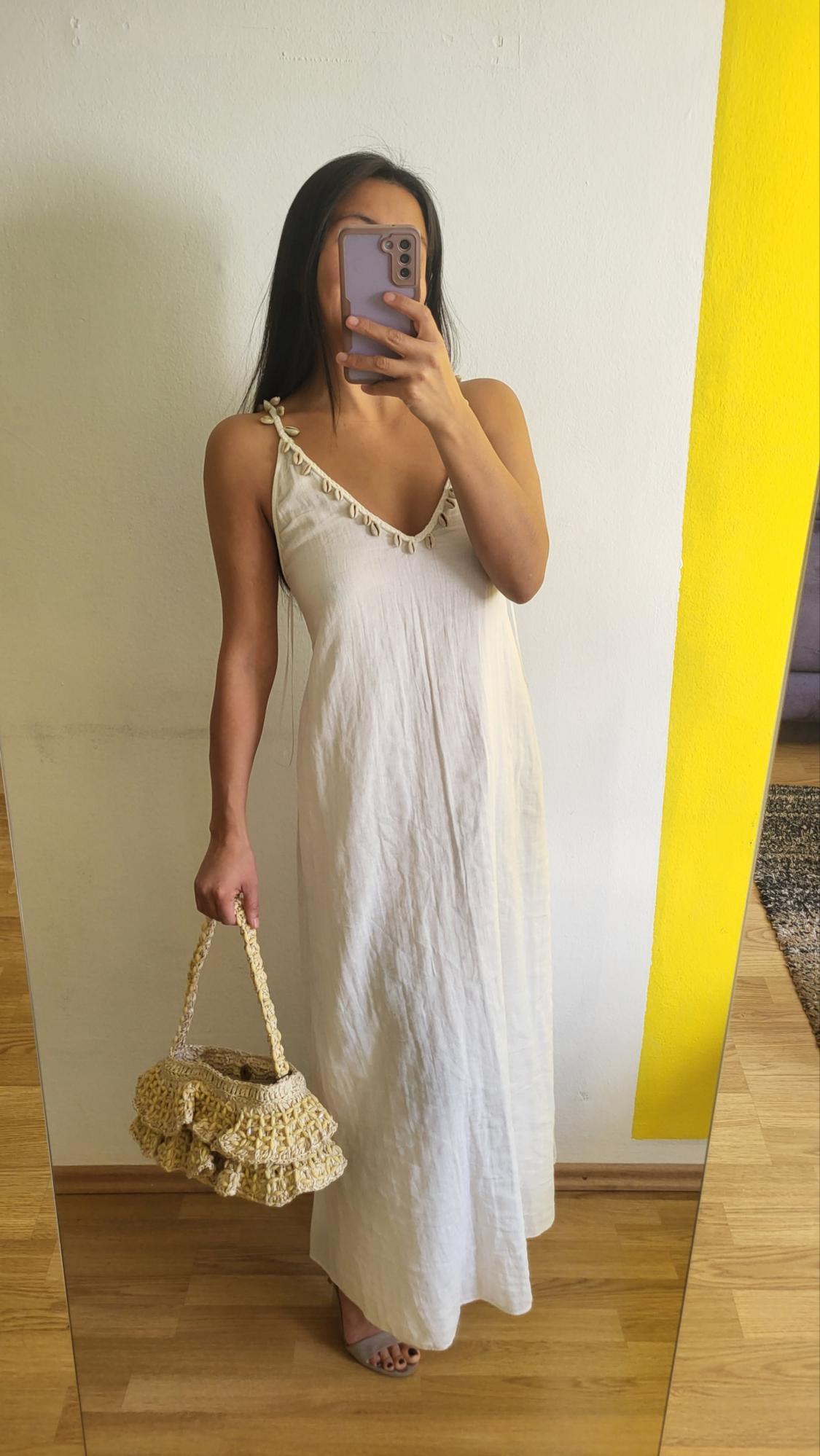 Woman in a long white dress and woven bag, taking a photo in front of a mirror. Background with white wall and yellow stripe.