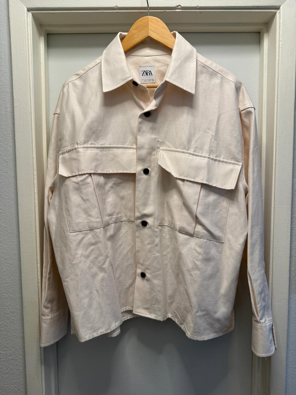 Beige long-sleeved shirt with black buttons and front pockets, hanging on a wooden hanger. Visible tag: Zara.
