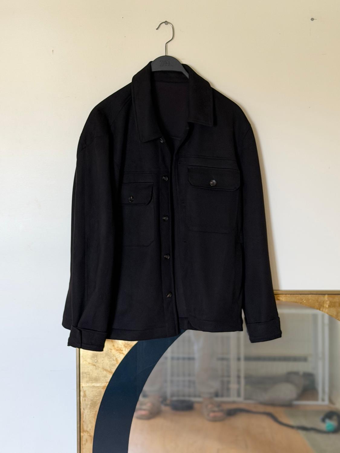 Black jacket hanging on a hanger against a light background. Reflection of a person in a mirror at the bottom.
