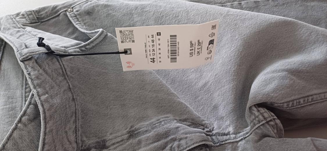 Grey trousers with a swing tag showing a barcode and size 44. Denim fabric on a light surface.