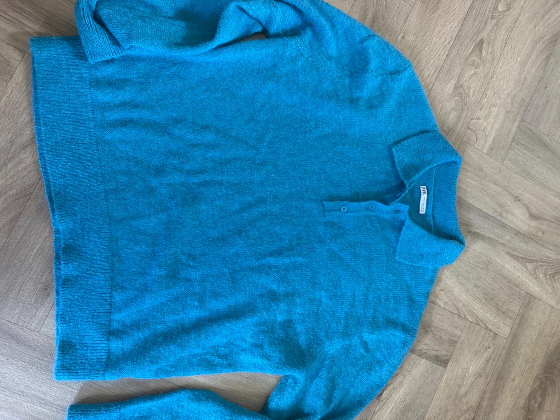 Blue knitted polo-neck jumper with buttons, laid out on a herringbone wooden floor. Inner label with the text ZARA.