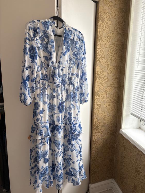 White dress with blue floral print hanging on a hanger. Background with gold wallpaper and window with blinds.