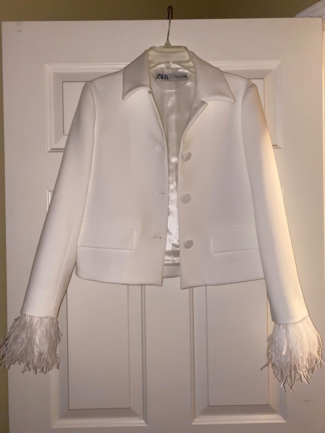White jacket with feathers on the cuffs, hanging on a door. Visible label with the text "ZARA".