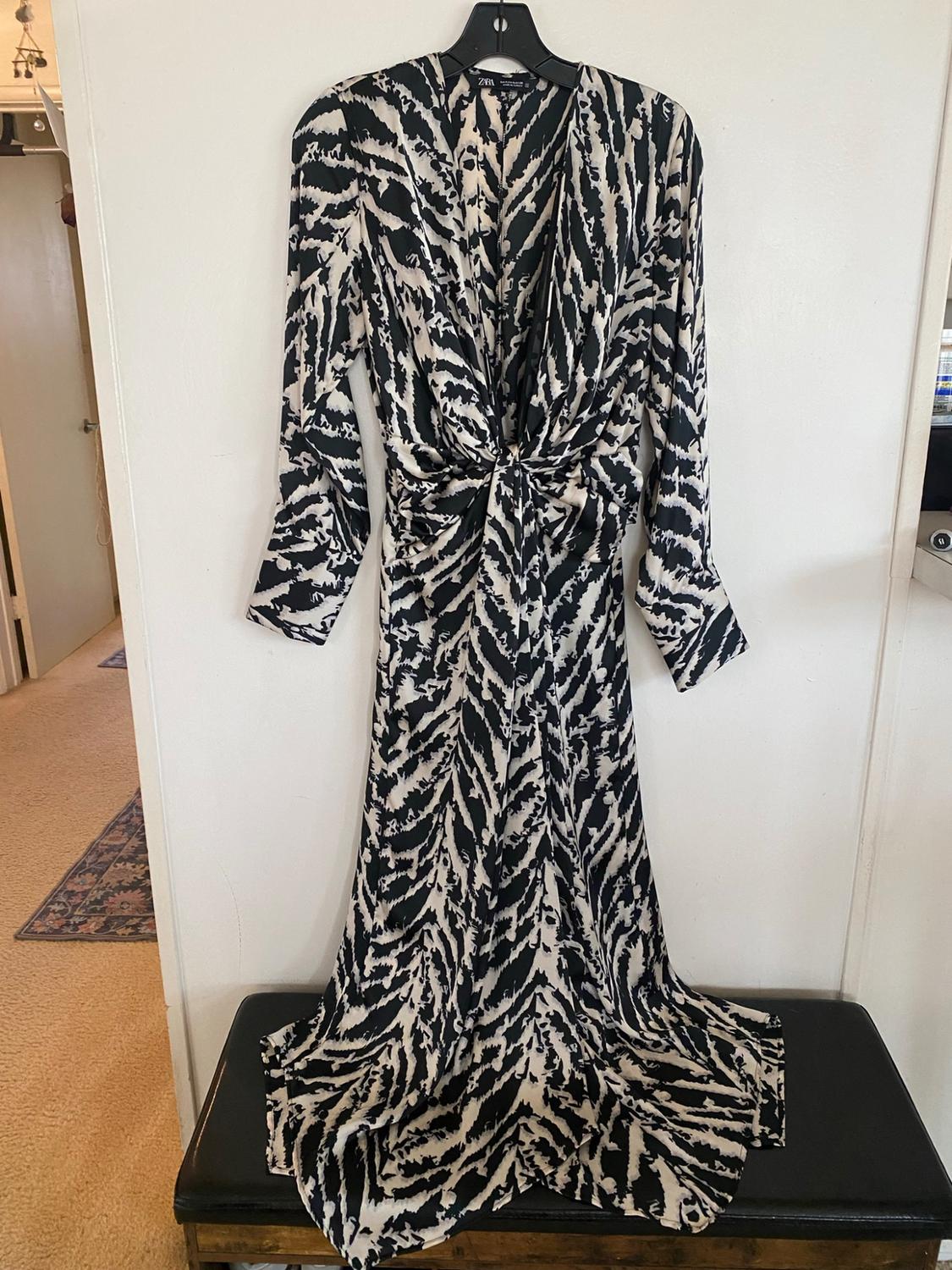 Black-and-white animal-print long dress, long sleeves, V-neck, and a front tie. ZARA label. Hanging on a hanger over a black bench.