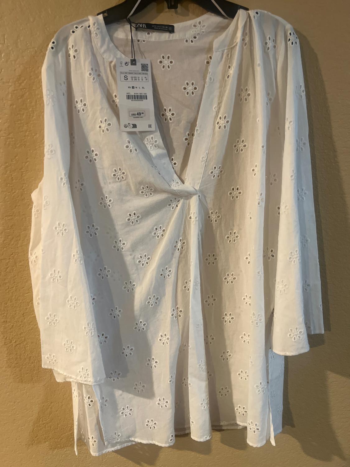 White, long-sleeved blouse with broderie anglaise floral embroidery, hanging on a hanger. Visible tag with price of 49.99.