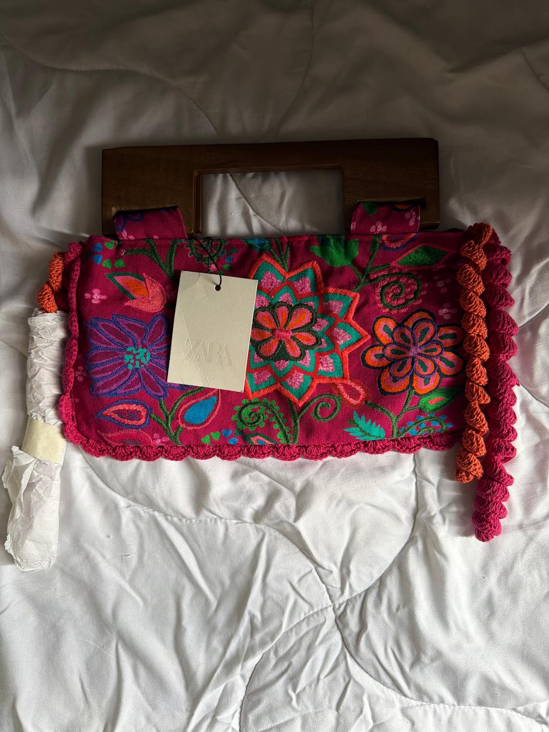 Colourful fabric bag with flowers and a wooden handle on a white bed. Label with "ZARA" text hanging from the bag.