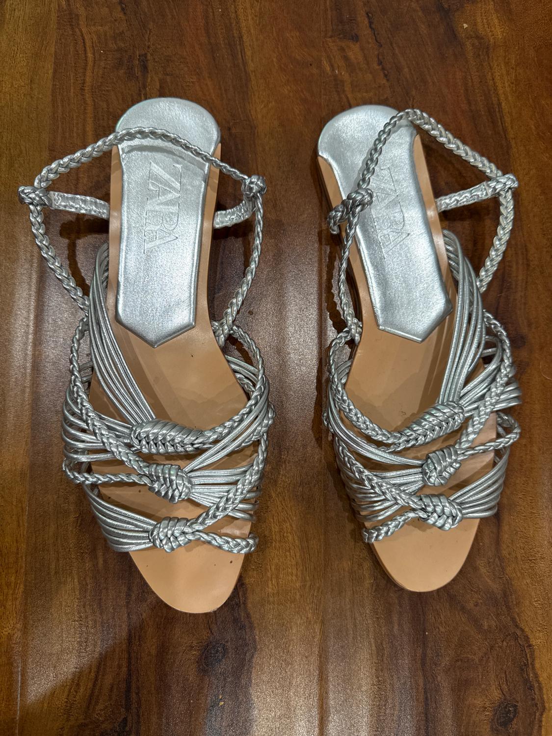 Silver sandals with braided straps on a wooden floor. The insole displays the "ZARA" brand.