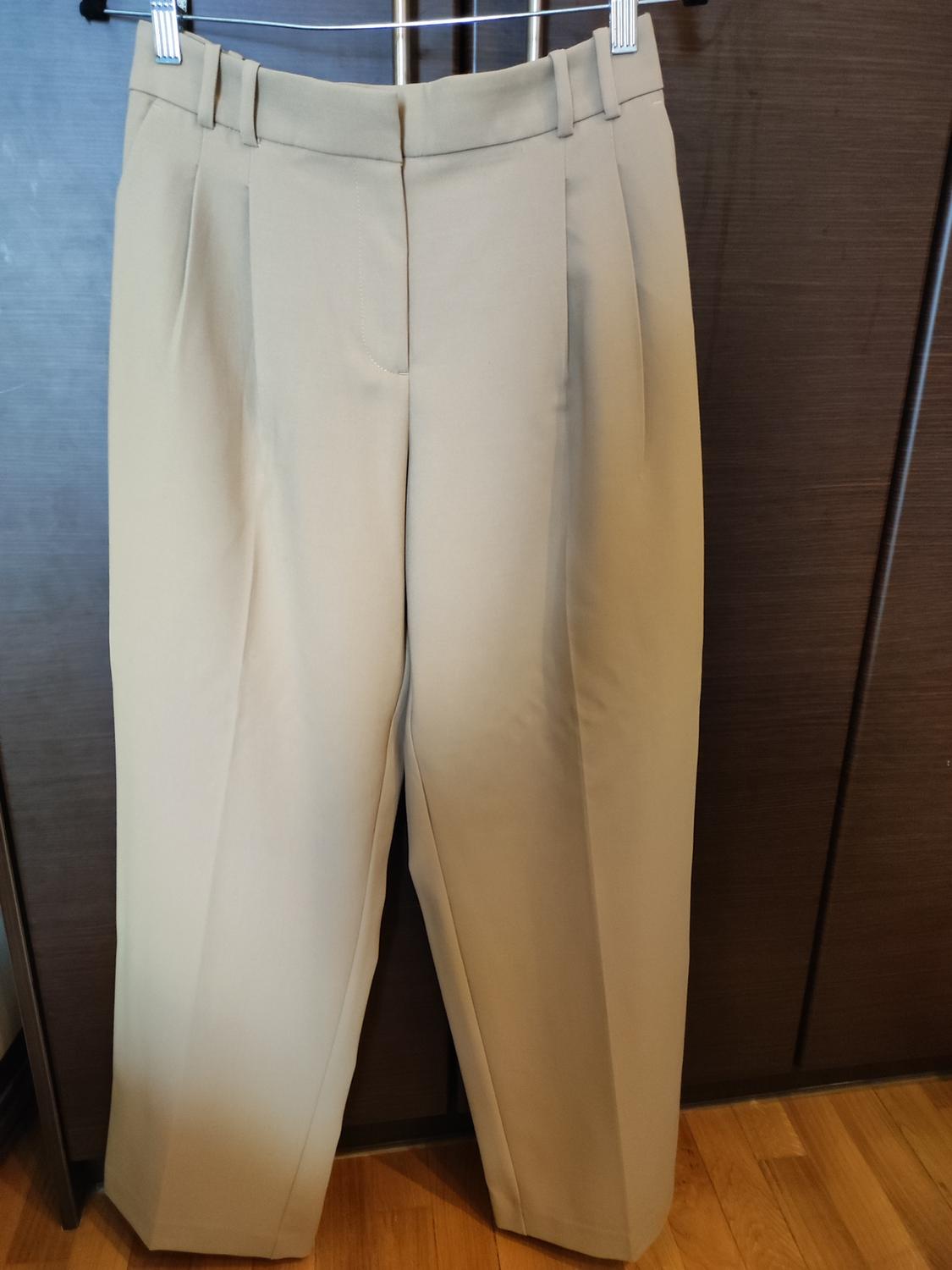 Beige trousers hanging on a hanger against a dark wooden background. Wooden floor visible at the bottom.