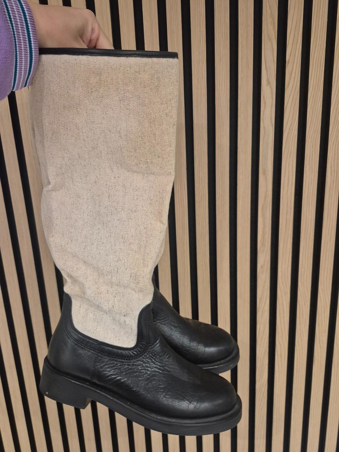 A pair of black leather and beige fabric knee-high boots held by a hand in front of a background of vertical wooden slats.