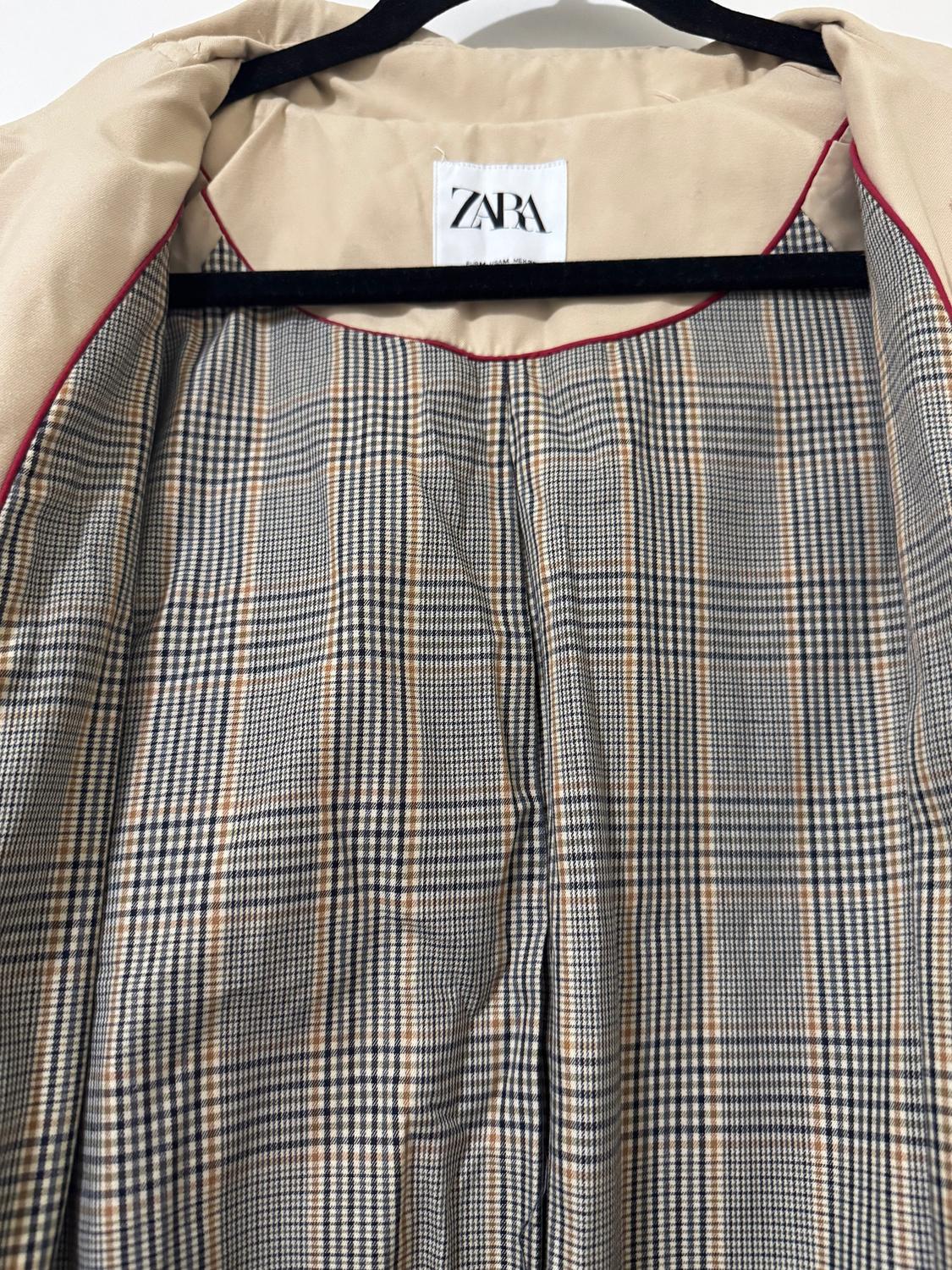 Beige coat with a plaid lining and red trim, hanging on a black hanger. Visible tag with the text "ZARA".