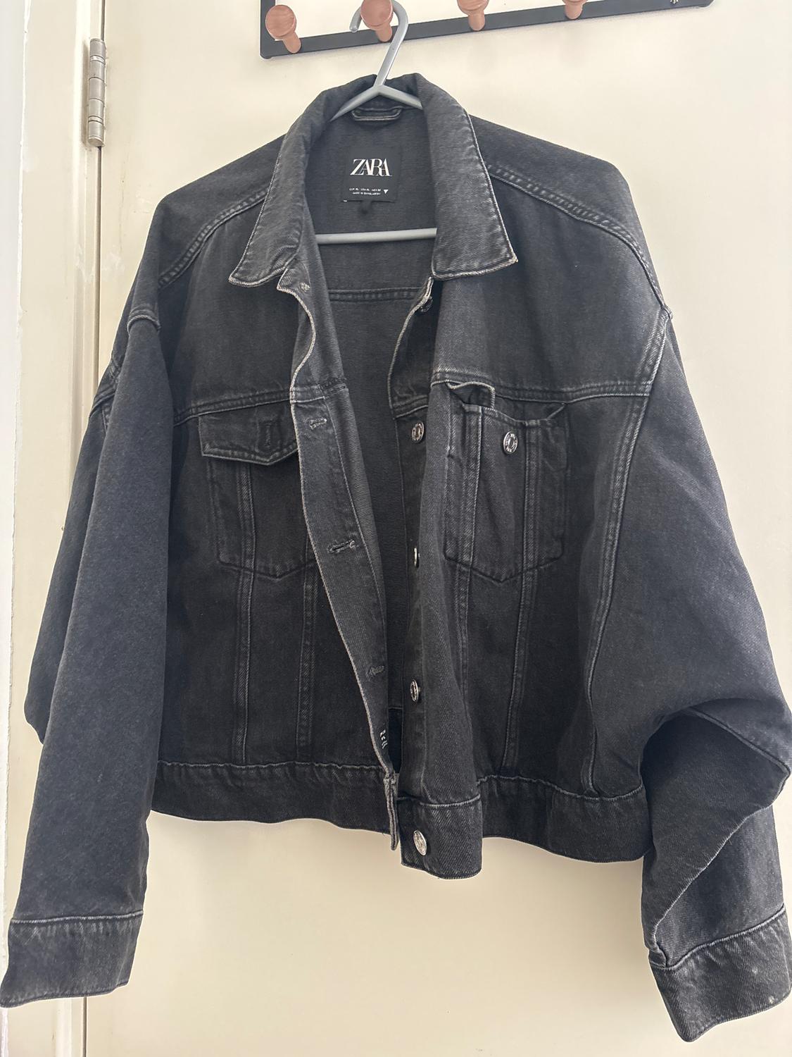 Black denim jacket hanging on a wooden hanger. Visible "ZARA" brand label on the collar.