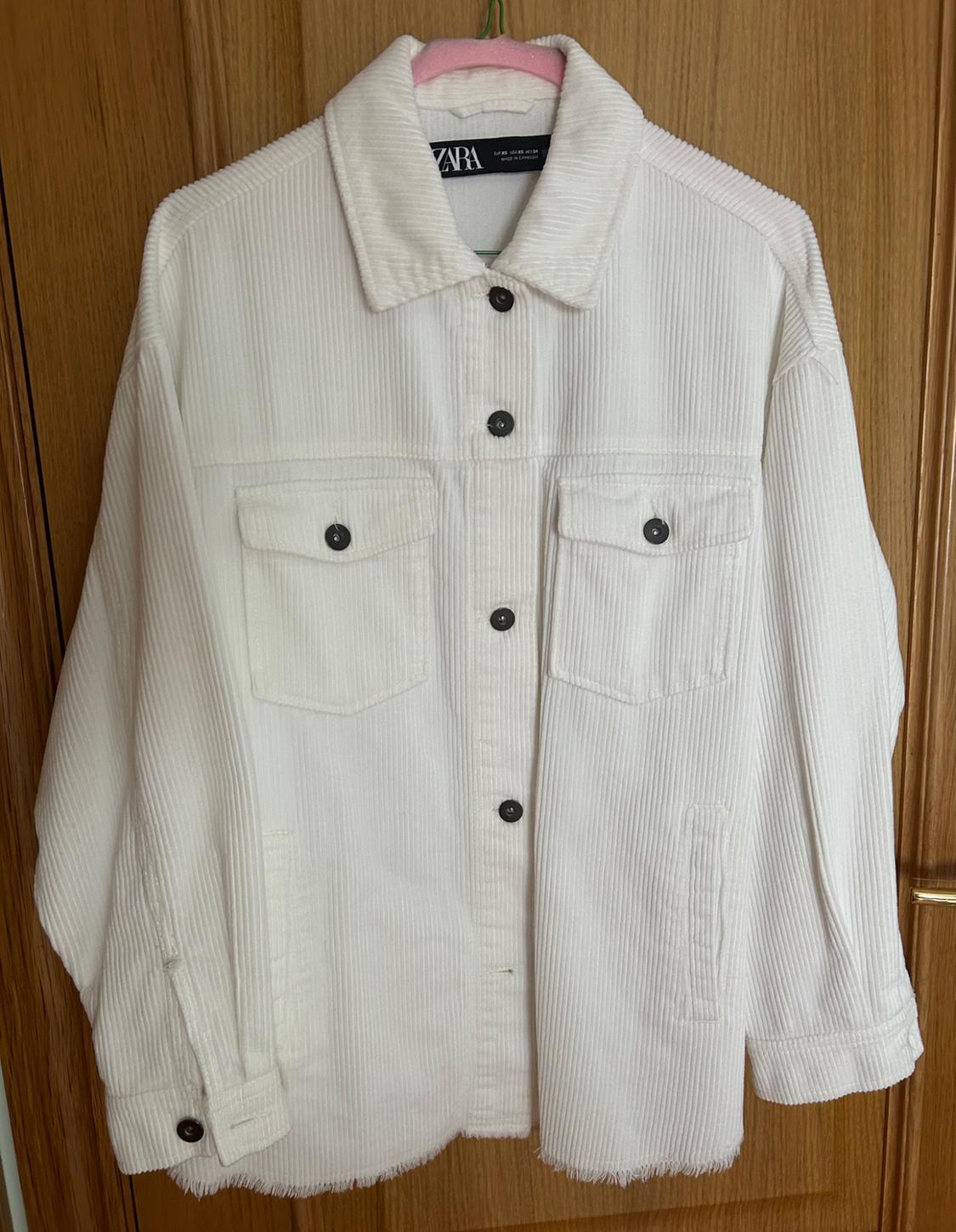 White corduroy jacket with black buttons and front pockets, hanging on a pink hanger in front of a wooden door.