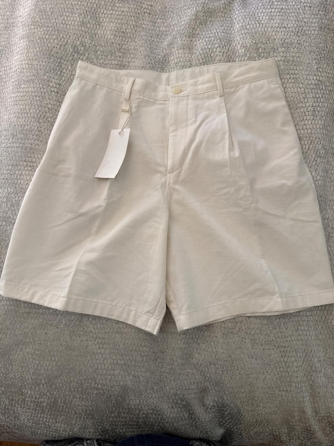 Beige tailored shorts with pleats and a button, on a textured grey fabric surface. Hanging tag with the text ZARA.