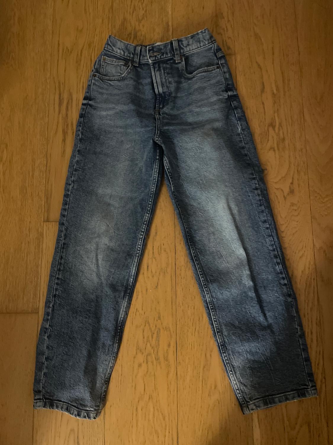 Worn blue jeans on a light wood floor. Straight leg, five pockets, and button and zip fastening.