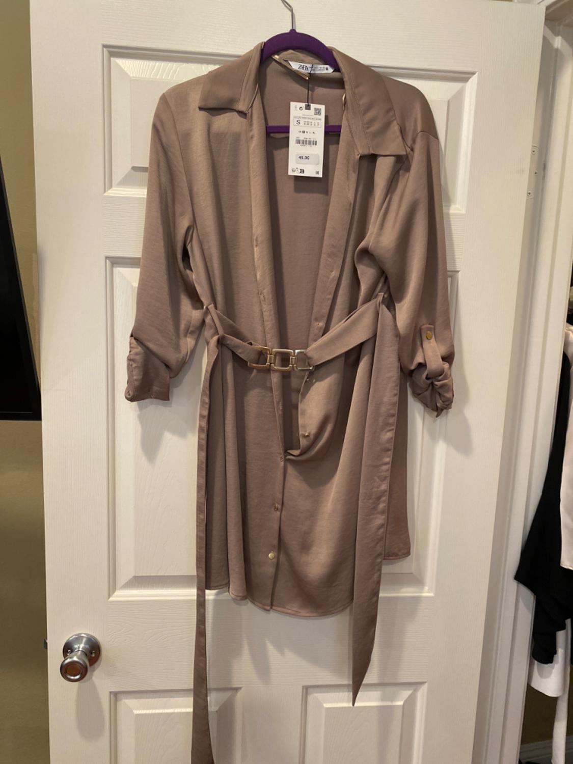 Beige shirt dress with adjustable belt and gold buttons, hanging on a purple hanger in front of a white door.