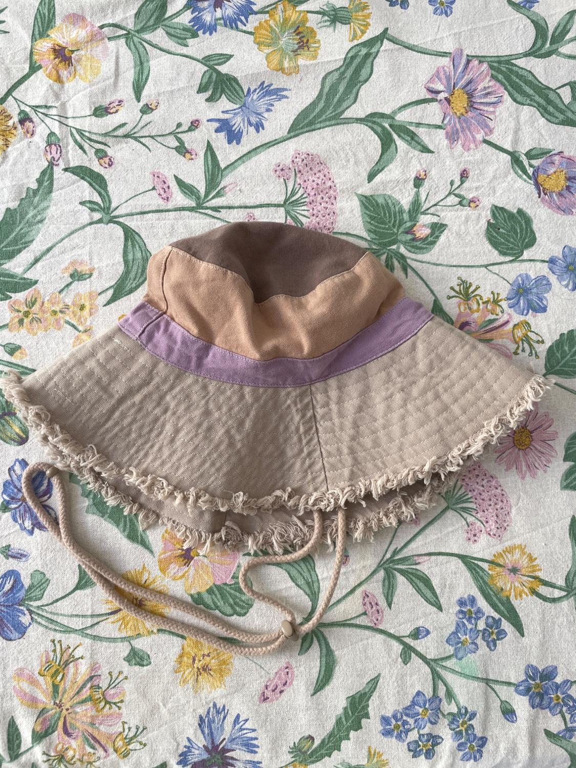 Fabric hat in shades of beige and lilac with a frayed edge, on a fabric background with a colourful floral print.