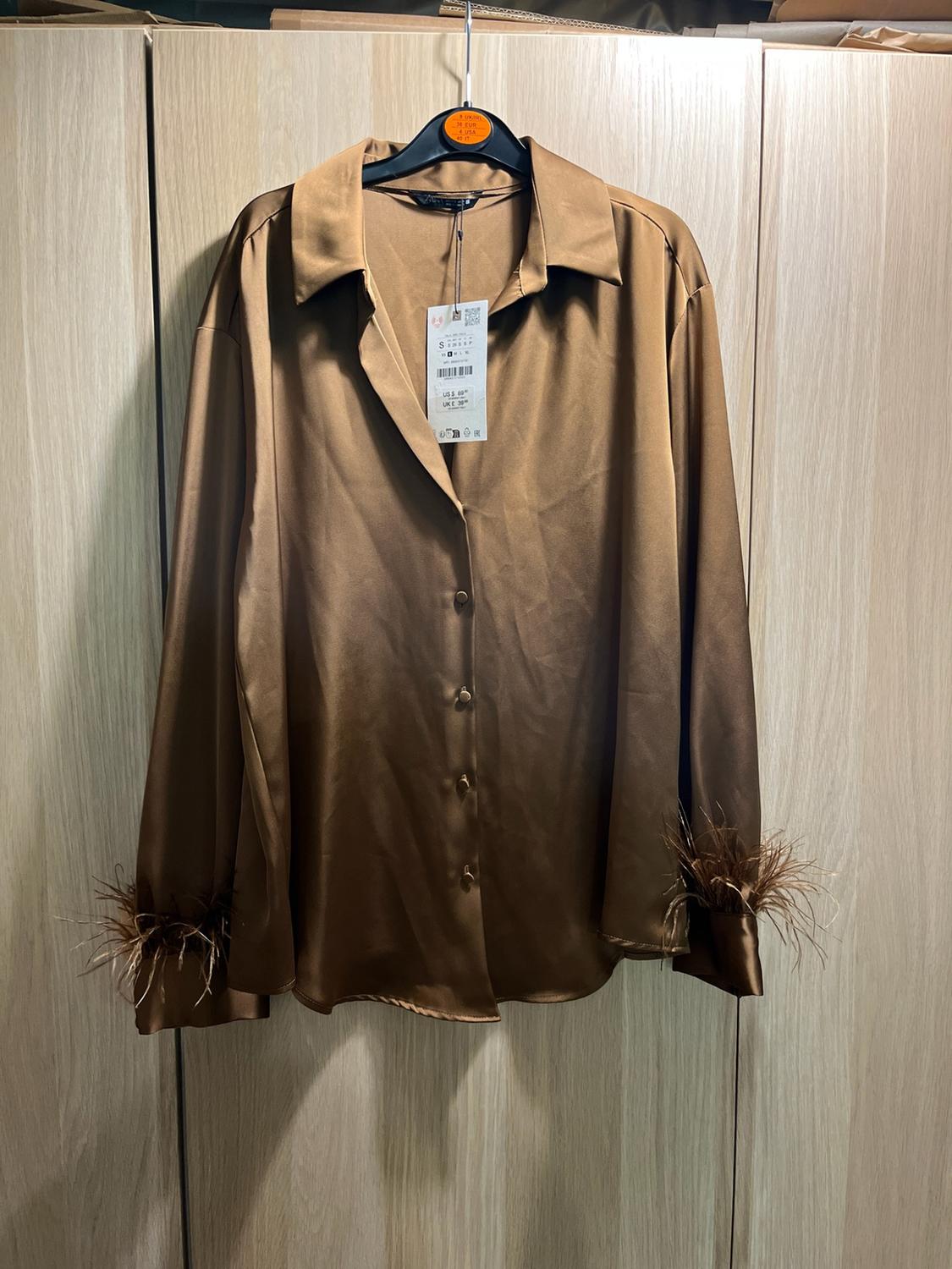 Brown satin long-sleeved shirt with buttons and feather detailing on the cuffs, hanging on a hanger in front of a wooden wardrobe.