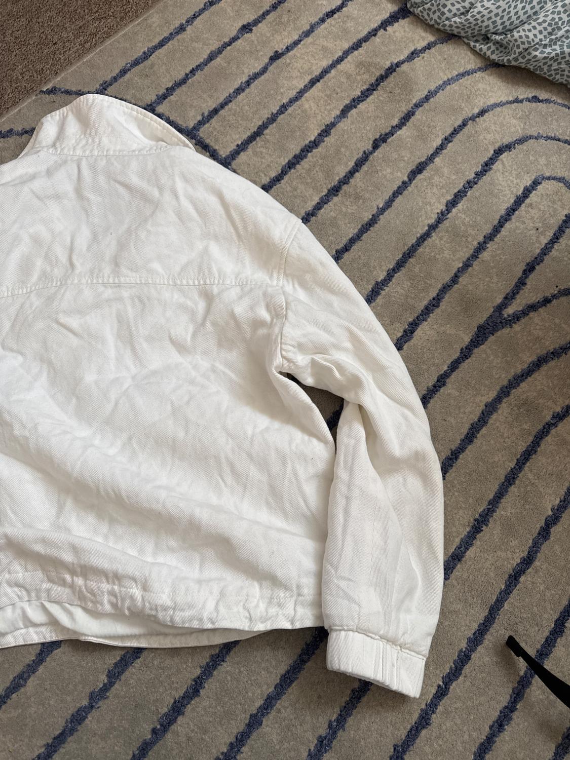 Creased white jacket seen from behind, on a beige rug with a blue curved line pattern. Relaxed composition.