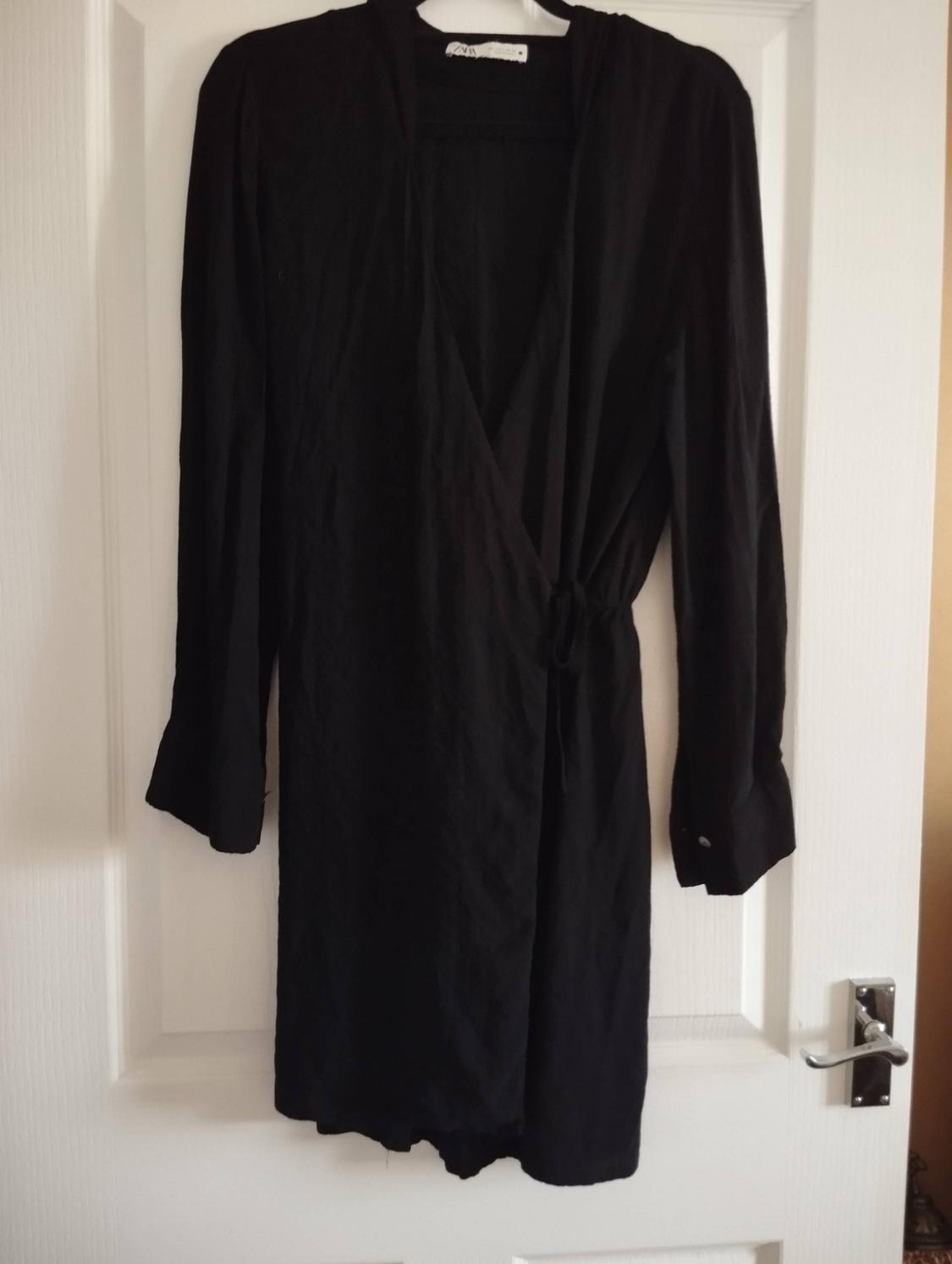Black long-sleeved wrap dress with a side tie, hanging on a hanger over a white door with a silver handle.