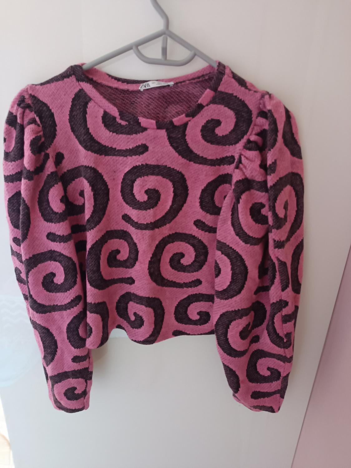 Pink jumper with a black spiral print, hanging on a grey hanger over a white door.