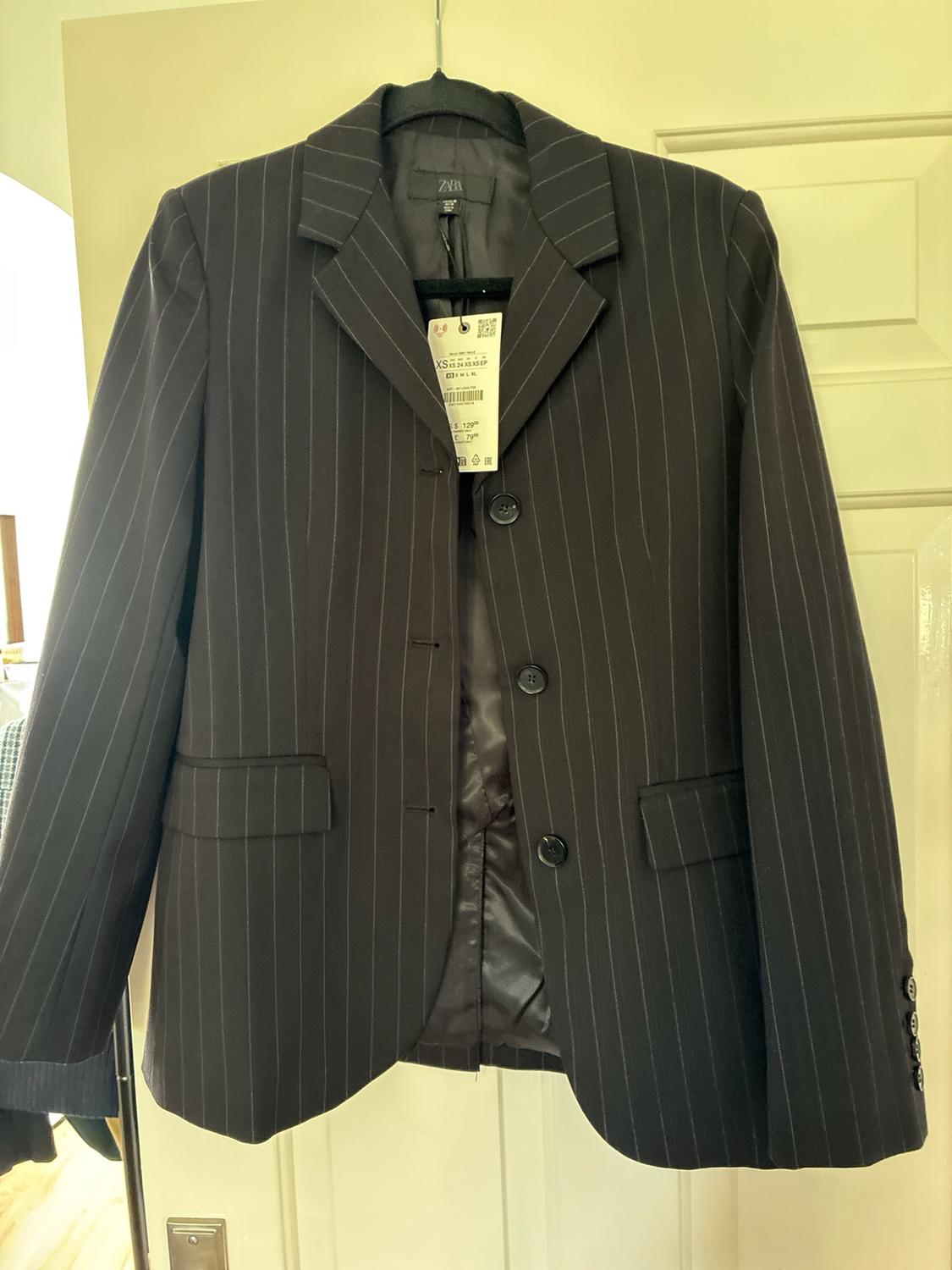 Black pinstriped blazer hanging on a door. It features three front
buttons. The inner label reads ZARA.