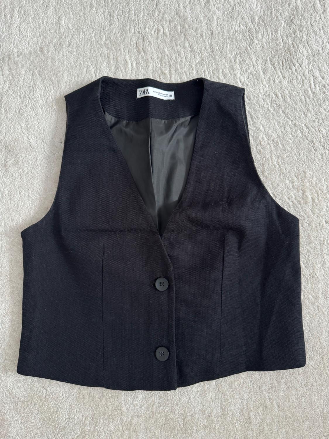 Women's black waistcoat with two buttons, on a light background. Visible label with text "ZARA" on the inside of the collar.