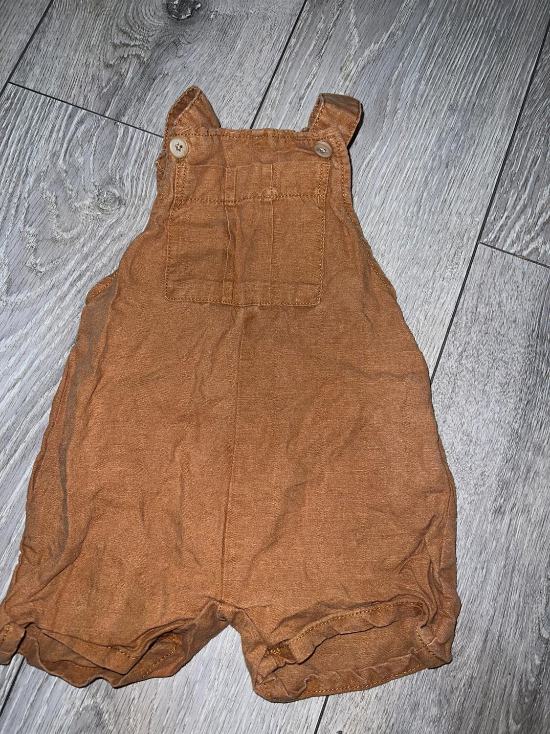 Brown baby romper with straps and wooden buttons, on grey wooden floor. Wrinkled fabric and front pocket.